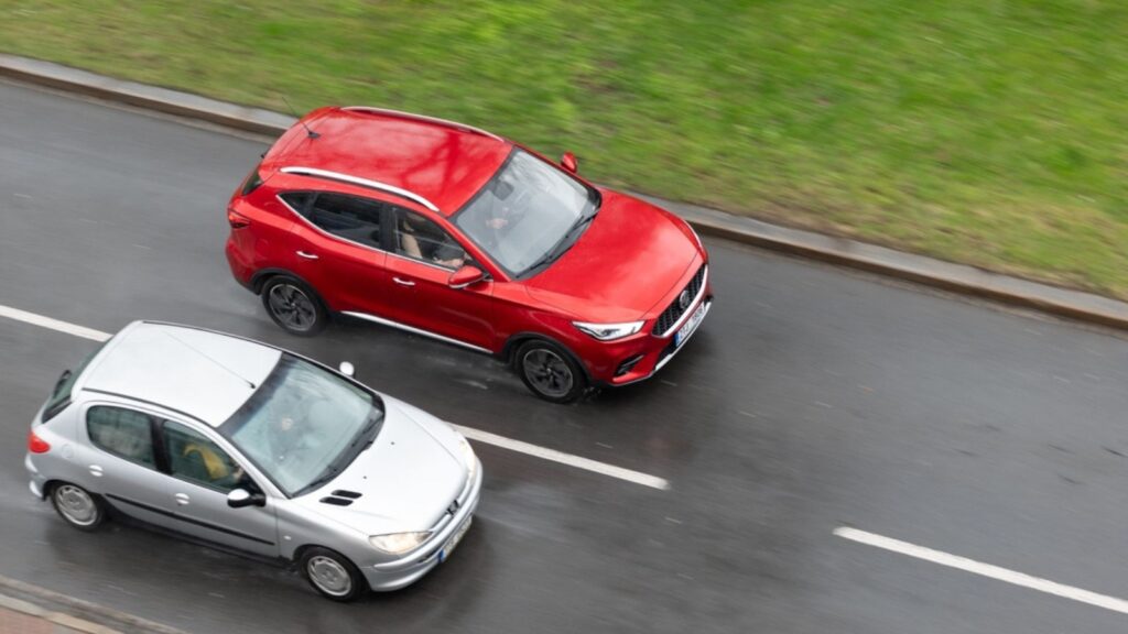 Red Chinese MG ZS crossover overtaking Peugeot 206 car in heavy rain on street