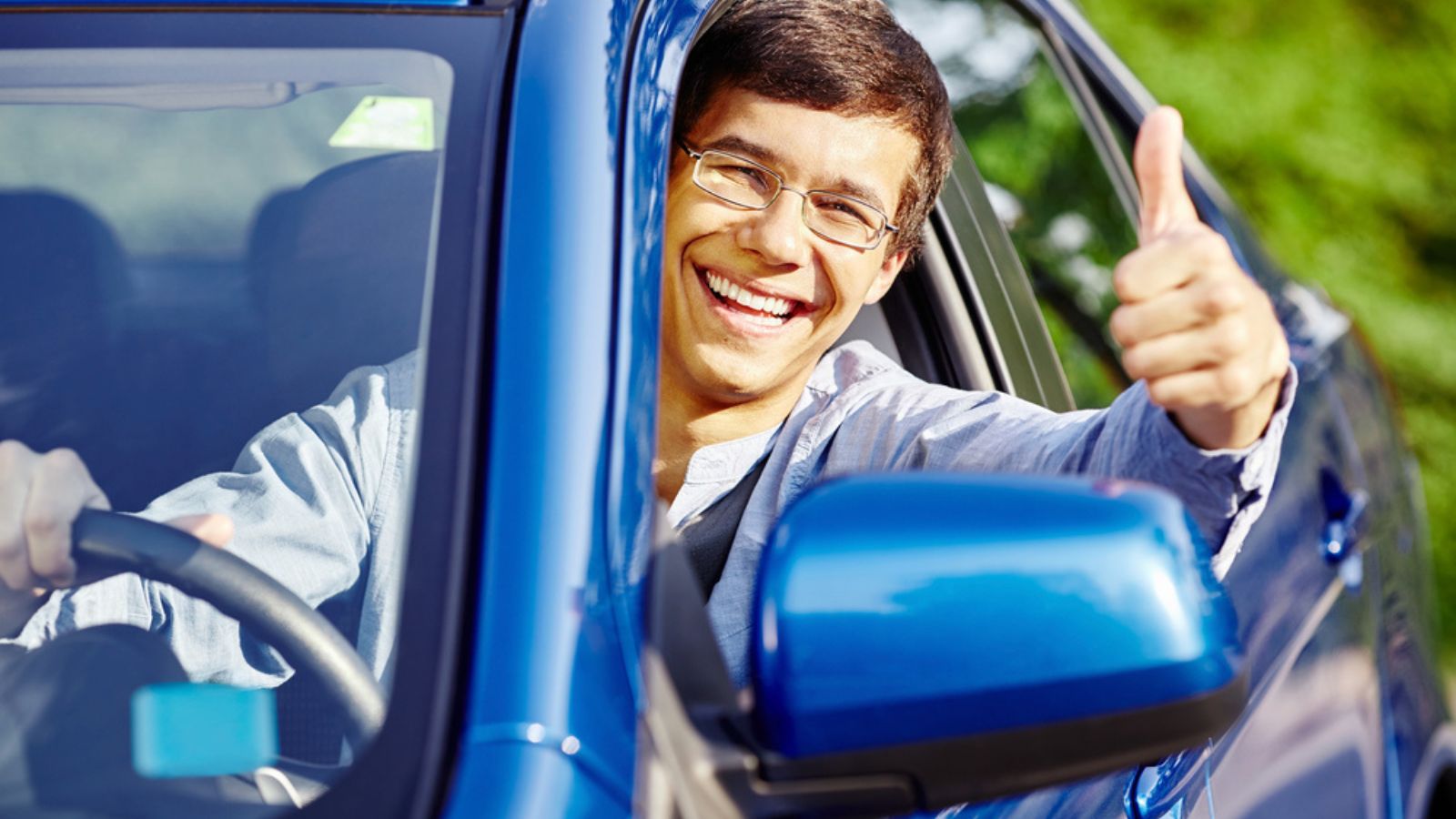 Young happy hispanic man wearing glasses sitting inside car, holding steering wheel, showing thumb up hand gesture through car window and laughing