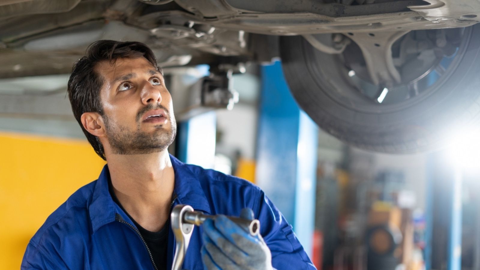 happy latin auto mechanic man checking tires , brakes under car with clipboard in garage cars service