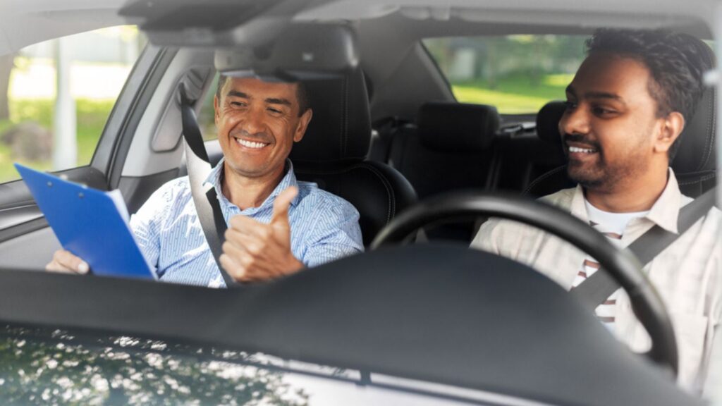 happy smiling indian man and driving school instructor with clipboard showing thumbs up in car
