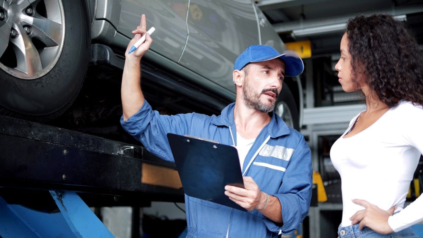 mechanic in blue apron gesturing at car talking to female client