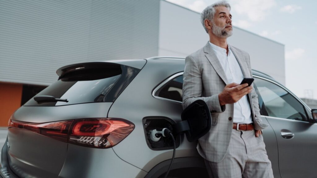 Businessman holding smartphone while charging car at electric vehicle charging station