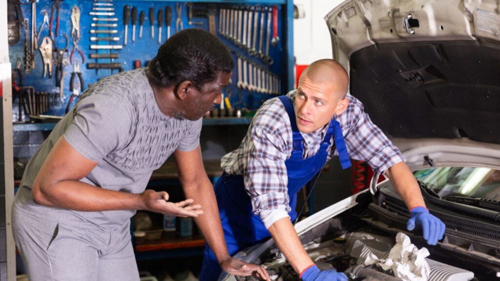 Worried man client talking with specialist in uniform about repairing his car engine at automotive workshop