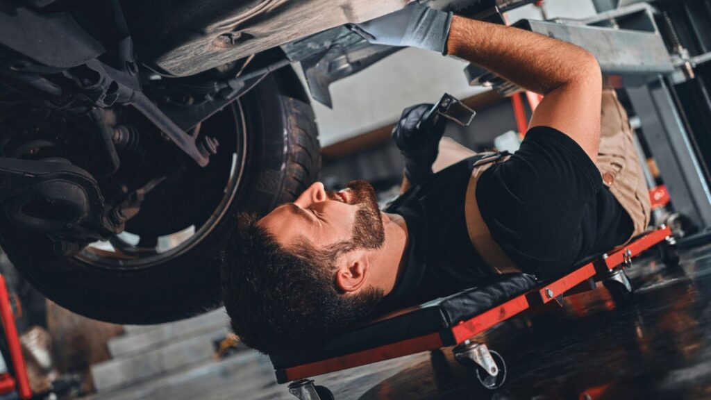 Bearded male technician in gloves lying on car creeper and using LED inspection lamp while working under automobile in garage