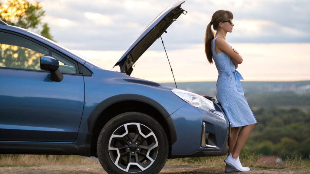 Young woman driver standing near a broken car with popped up hood waiting for help to arrive