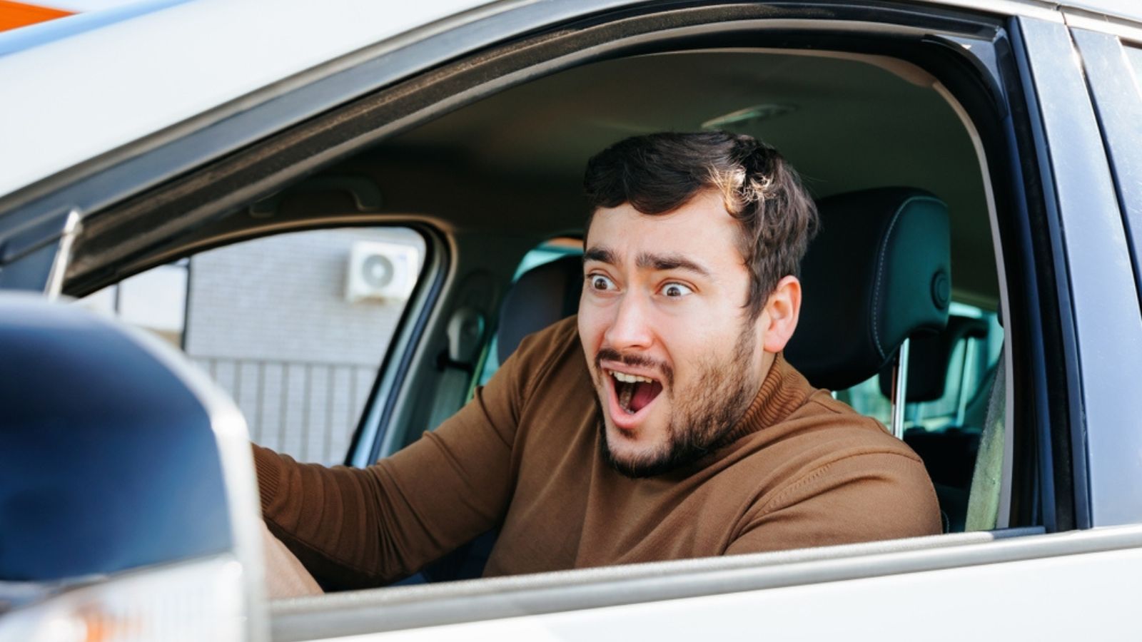 A frightened surprised young guy in a car behind the wheel screams with bulging eyes and looks ahead