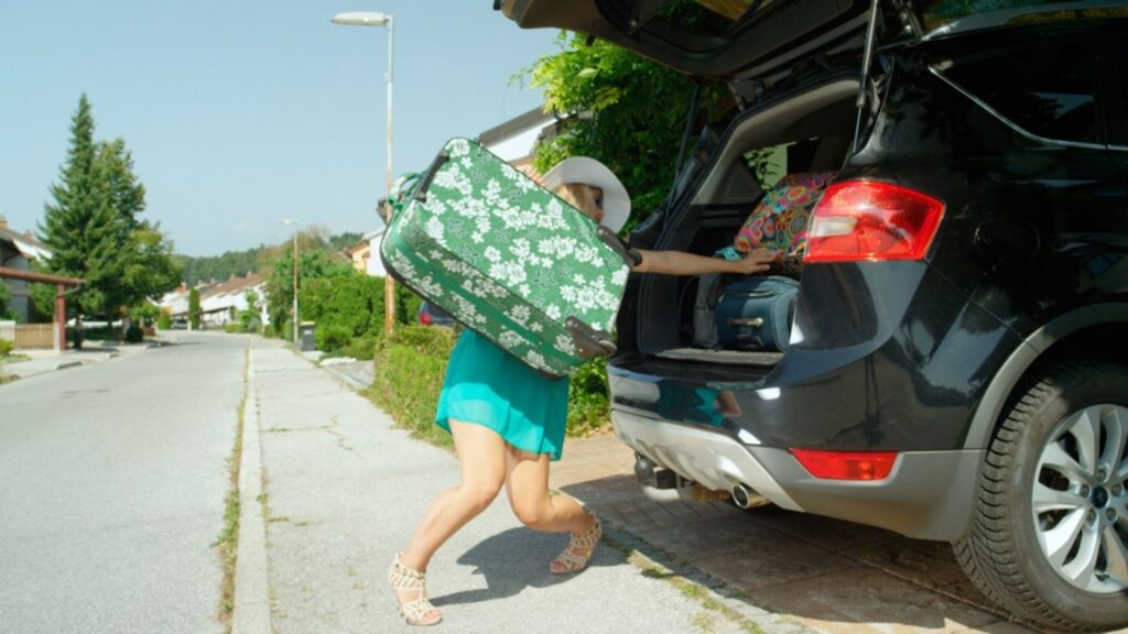 Blonde girl lifting a heavy suitcase while trying to keep all her other luggage from falling out of the car trunk 
