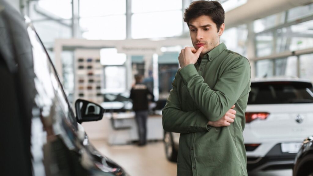 Pensive young guy thinking, making choice, having doubt about buying new car at dealership