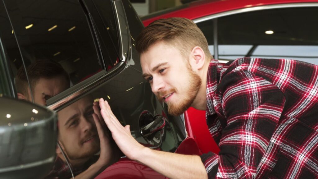 Cheerful young handsome man smiling touching a new auto at the dealership examining car paint